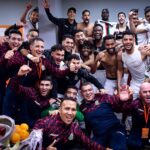 Large group of soccer players and staff celebrate in a locker room posing for a group photo with a fruit bowl in the foreground  trybalnews