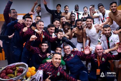 Large group of soccer players and staff celebrate in a locker room, posing for a group photo with a fruit bowl in the foreground.