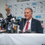 Carlos Queiroz in a dark suit and red tie speaks at a press conference pointing with one finger microphones crowded on the table behind him and a sponsor backdrop visible  trybalnews