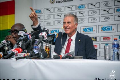 Carlos Queiroz in a dark suit and red tie speaks at a press conference, pointing with one finger, microphones crowded on the table behind him and a sponsor backdrop visible.