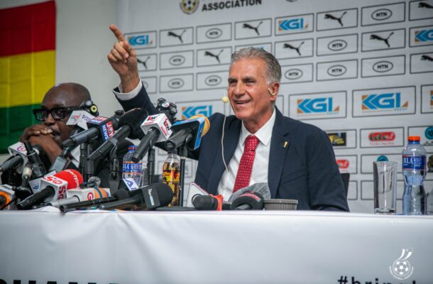 Carlos Queiroz in a dark suit and red tie speaks at a press conference pointing with one finger microphones crowded on the table behind him and a sponsor backdrop visible  trybalnews