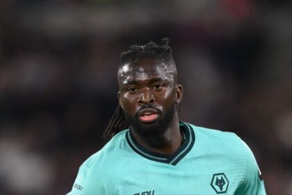 Football player in a teal Wolverhampton kit on the pitch, sweat glistening, with 'DEBET' sponsor across the chest and a wolf crest visible on the jersey.