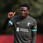 Football player in a dark gray Liverpool training top raising his right hand sweaty and focused on a practice field  trybalnews