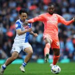 Two soccer players contest a ball during a match one in blue and white kit the other in orange with a crowded stadium in the background  trybalnews