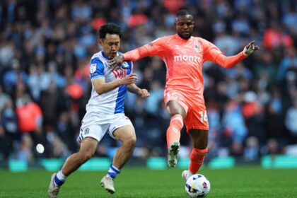 Two soccer players contest a ball during a match: one in blue-and-white kit, the other in orange, with a crowded stadium in the background.