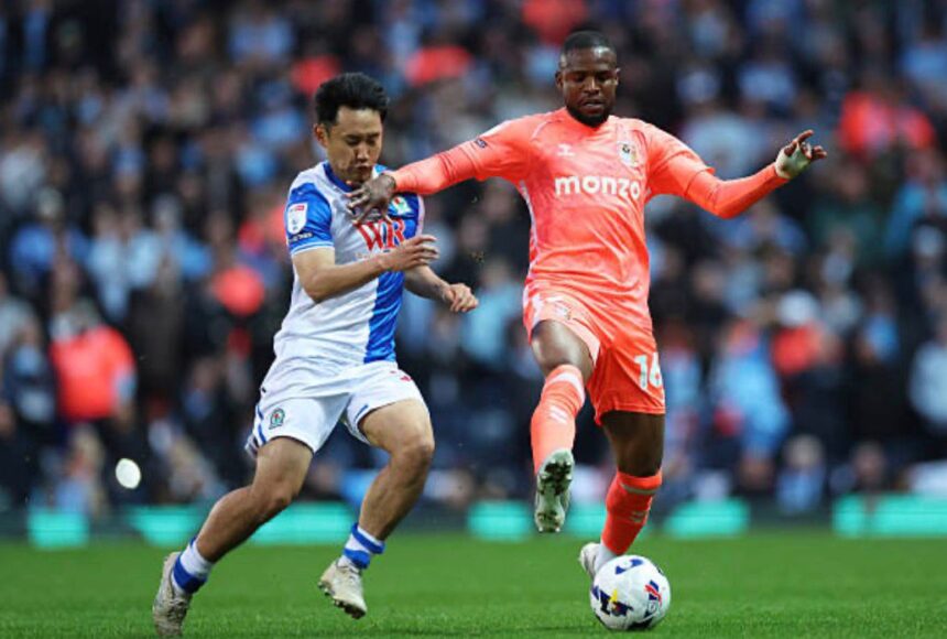 Two soccer players contest a ball during a match one in blue and white kit the other in orange with a crowded stadium in the background  trybalnews