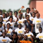 Group of young men in white Promoted T shirts posing together cheering with fists raised outdoors The team photo conveys celebration and camaraderie  trybalnews