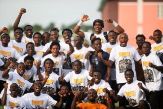 Group of young men in white Promoted T shirts posing together cheering with fists raised outdoors The team photo conveys celebration and camaraderie  trybalnews
