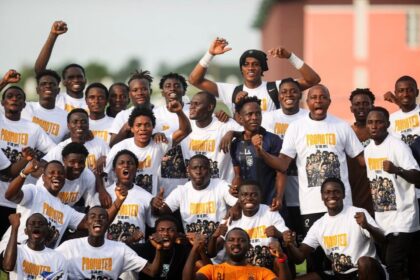 Group of young men in white 'Promoted' T-shirts posing together, cheering with fists raised outdoors. The team photo conveys celebration and camaraderie.