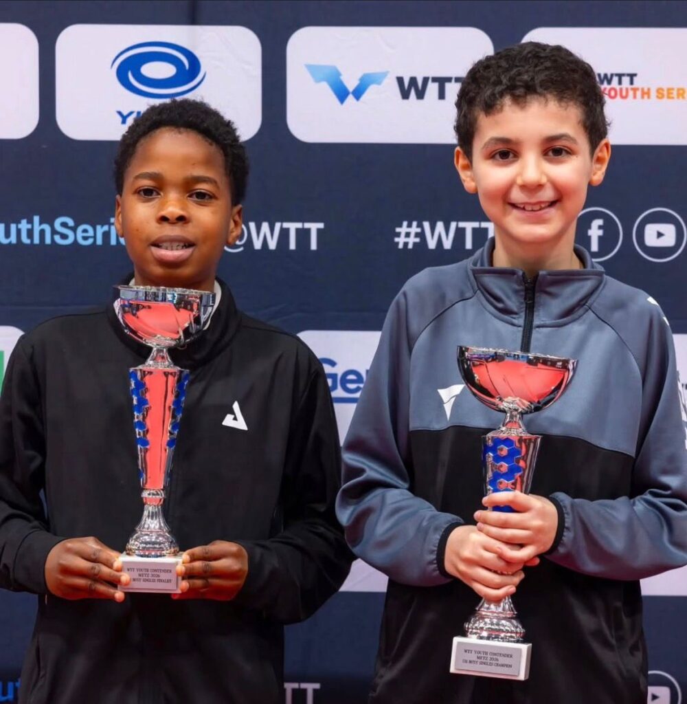 Two boys stand side by side holding shiny trophies and smiling at a awards backdrop with logos behind them  trybalnews