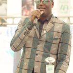 Man in a teal brown patterned suit wearing sunglasses speaking into a microphone at a podium during an event with event banners in the background  trybalnews