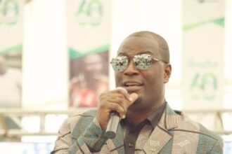 Man in a teal brown patterned suit wearing sunglasses speaking into a microphone at a podium during an event with event banners in the background  trybalnews