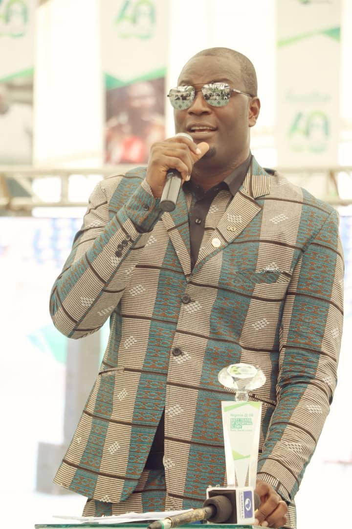 Man in a teal brown patterned suit wearing sunglasses speaking into a microphone at a podium during an event with event banners in the background  trybalnews