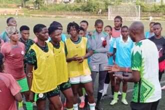 Coach speaking to a group of young soccer players wearing yellow bibs on a turf field.