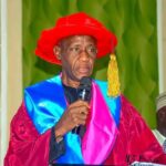 A man in a bright red graduation cap and maroon gown speaks into a handheld microphone at a ceremony with a blue sash over pink trim behind him informative  trybalnews