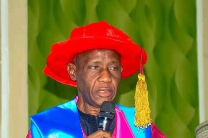 A man in a bright red graduation cap and maroon gown speaks into a handheld microphone at a ceremony, with a blue sash over pink trim behind him (informative).