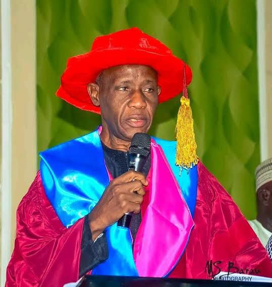A man in a bright red graduation cap and maroon gown speaks into a handheld microphone at a ceremony with a blue sash over pink trim behind him informative  trybalnews