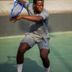 A male tennis player in gray athletic wear swings a blue racket during a match on an outdoor court with a focused expression from the side angle  trybalnews