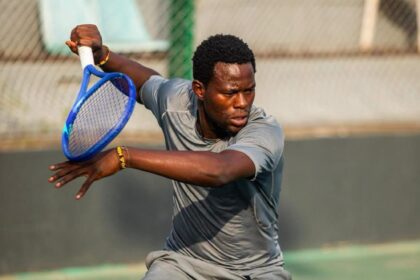 A male tennis player in gray athletic wear swings a blue racket during a match on an outdoor court with a focused expression from the side angle.