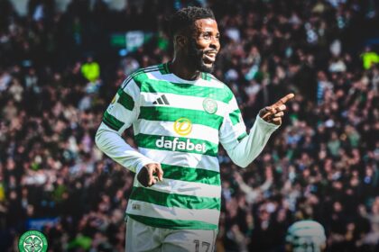 Celtic FC player in green-and-white hoops celebrating, pointing with his right hand while the crowded stadium cheers behind him.