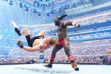 Two muscular wrestlers clash in a packed arena; one fighter flips backward as the other delivers a kick.