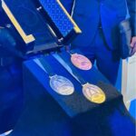 Three circular medals silver bronze gold with blue ribbons laid on a dark blue display cloth a presentation case behind them  trybalnews