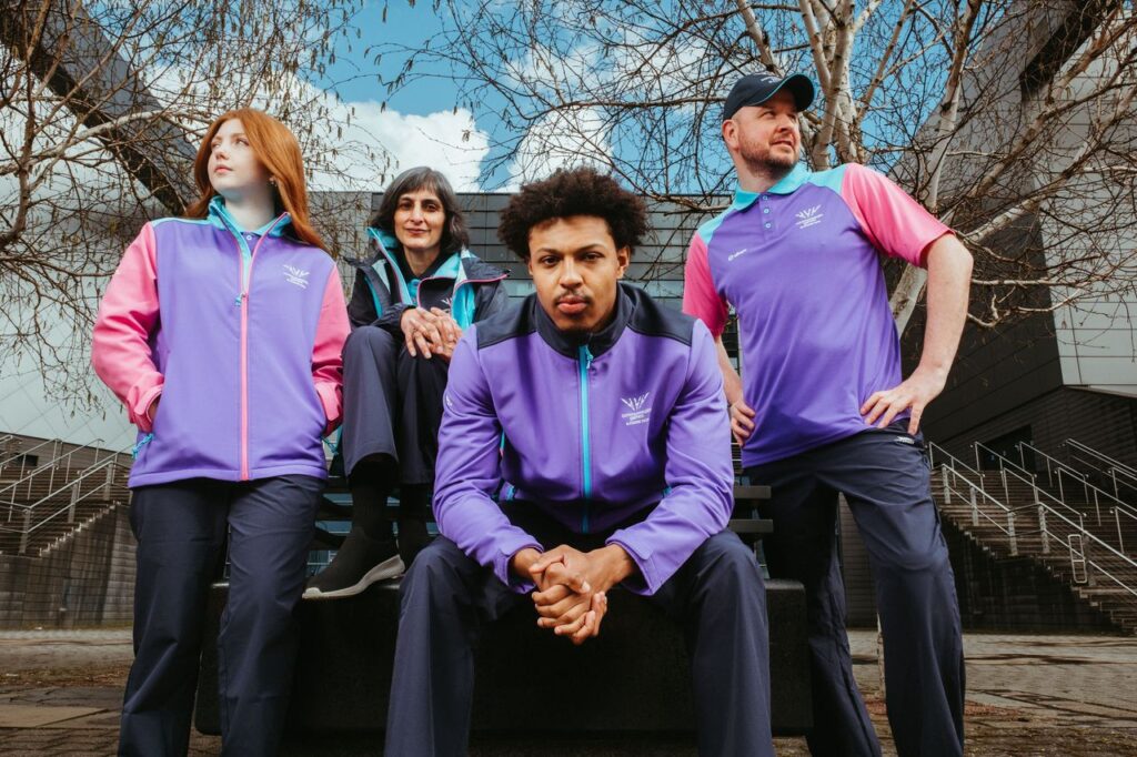 Group of four people in purple and pink athletic jackets posing outdoors on a bench in front of a modern building  trybalnews