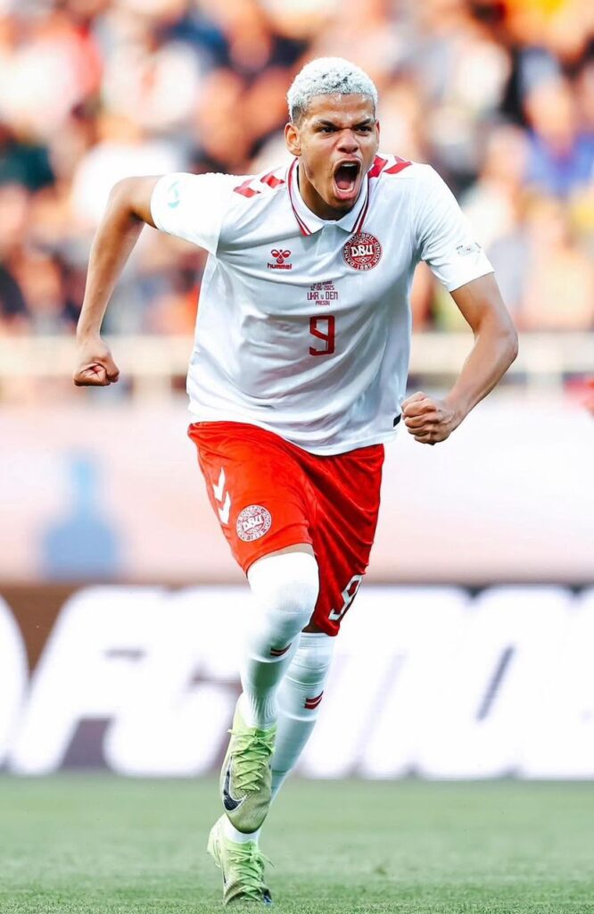 Soccer player in white and red kit sprinting and shouting in celebration on a green field with a blurred crowd behind him  trybalnews