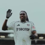 Soccer player in a white Fulham kit wearing black gloves raising his left hand and smiling on the pitch  trybalnews