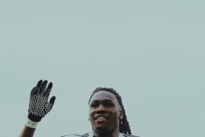 Soccer player in a white Fulham kit, wearing black gloves, raising his left hand and smiling on the pitch.
