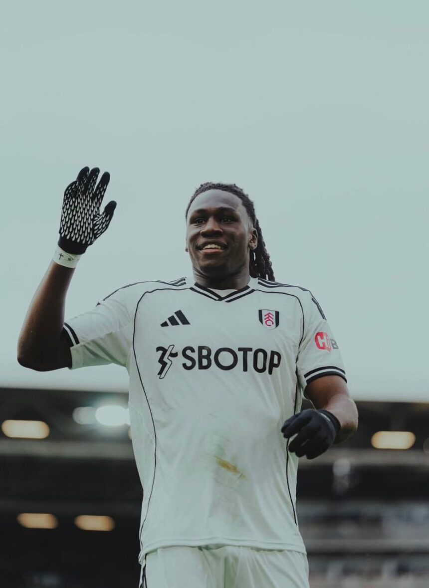 Soccer player in a white Fulham kit wearing black gloves raising his left hand and smiling on the pitch  trybalnews