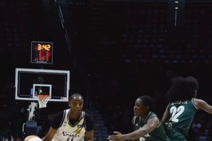 Female basketball player in white jersey dribbles toward the basket, pursued by green-jersey defenders on a polished court.