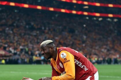Soccer player in a red Galatasaray jersey with the number 45 and orange sleeves bends over to adjust his bandaged right hand on a green field, with a cheering crowd in the stadium behind him.