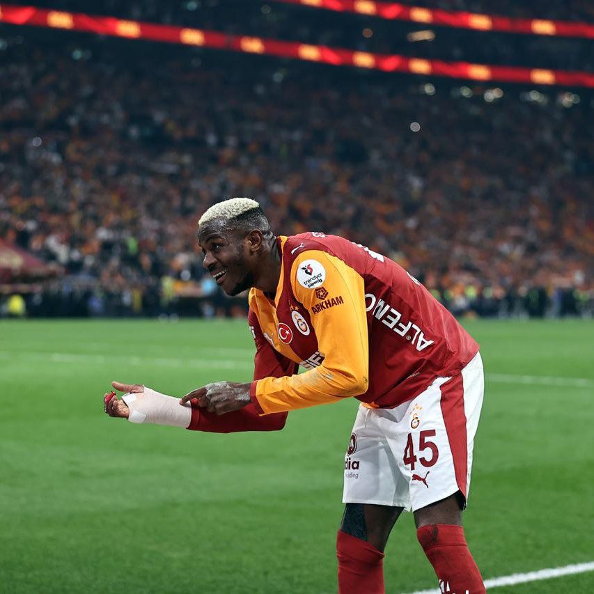 Soccer player in a red Galatasaray jersey with the number 45 and orange sleeves bends over to adjust his bandaged right hand on a green field with a cheering crowd in the stadium behind him  trybalnews