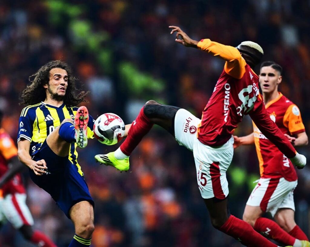 WhatsApp Image 20260426 at 201750 Trybal News Two soccer players contest the ball mid air during a match one in yellow and navy stripes and the other in red and white kit with a crowd in the background trybalnews