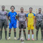 Five young men in soccer kits stand in a line on a field with a ball at their feet ready for kick off Behind them is a stadium wall  trybalnews