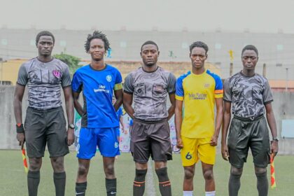 Five young men in soccer kits stand in a line on a field with a ball at their feet, ready for kick-off. Behind them is a stadium wall.