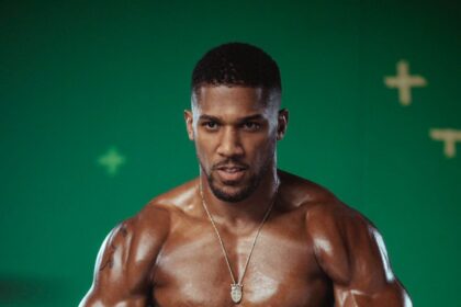 Muscular male boxer in white gloves and shorts, fists raised, standing in front of a green backdrop.
