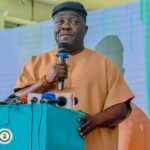 Man in brown traditional attire speaks into a microphone at a turquoise podium with multiple microphones in front of him during a press event  trybalnews