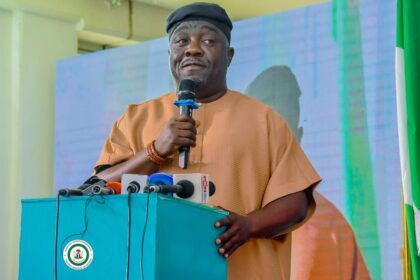 Man in brown traditional attire speaks into a microphone at a turquoise podium with multiple microphones in front of him, during a press event.