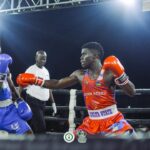 Two boxers exchange punches in a ring during a night match red team delta blue trunks and a referee behind them under bright lights audience out of frame  trybalnews