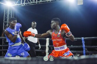 Two boxers exchange punches in a ring during a night match: red team delta, blue trunks, and a referee behind them under bright lights, audience out of frame.