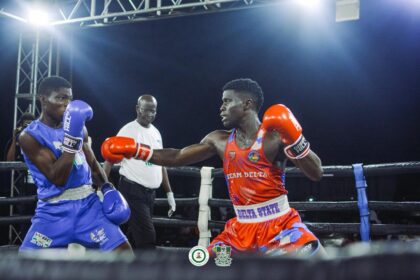 Two boxers exchange punches in a ring during a night match: red team delta, blue trunks, and a referee behind them under bright lights, audience out of frame.