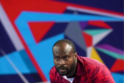 Nigerian table tennis player in a maroon GEWO jersey crouches at the net, focused during a match.
