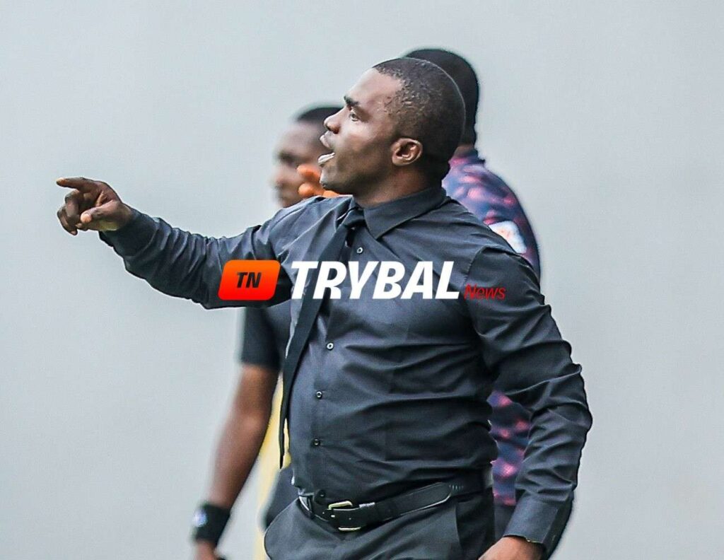 Dark skinned man in a black button down shirt points to the left during a discussion with a TRYBAL News watermark across his torso  trybalnews