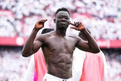 Athlete in a stadium celebration, shirtless, with hands raised near his ears as the crowd cheers behind him.