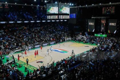 Packed basketball arena during a live game; players on court, sponsor banners along the sidelines.