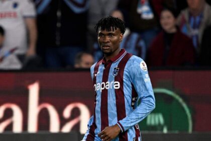 Professional footballer in blue and maroon striped jersey running on a soccer field with spectators in the stands behind him.