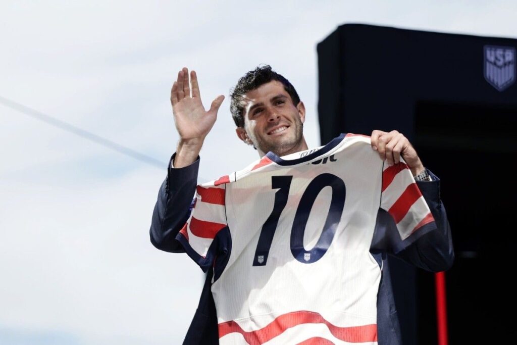 ChristianPulisic Trybal News Male athlete on a stage waving with one hand while holding up a white jersey with the number 10 US Soccer branding visible in the background trybalnews