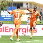 Orange clad soccer players celebrate a goal as one raises a fist while a teammate approaches a purple clad goalkeeper kneels on the turf nearby  trybalnews